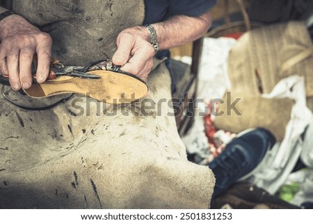 Close-up of the hands of an old master shoemaker in a leather apron using pliers to repair a shoe. In his messy workshop. Concept: Shoe repair circular economy and ecology against planned obsolescence
