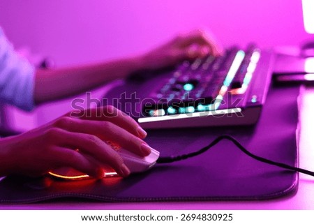 Close-up of hands playing on a computer keyboard and mouse in pink LED lighting. Modern gaming setup with neon glow and cyber aesthetic.