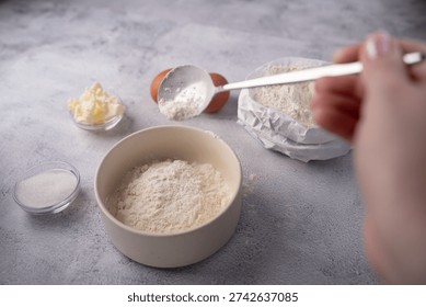 close-up of hands preparing baking ingredients on a textured grey countertop: a ceramic bowl and paper-wrapped bag of flour, a spoon sprinkling flour, eggs, butter and sugar in glass bowls, soft natur