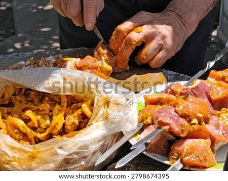 Close-up of hands preparing marinated meat for grilling outdoors. Pieces of raw meat coated with seasoning and marinade are being cut and threaded onto metal skewers. Plastic bag with marinated meat, plate, spices, sunlight and shadows visible. Food preparation scene before barbecue cooking.