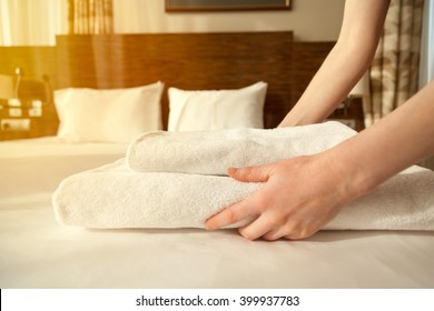 Close-up Of Hands Putting Stack Of Fresh White Bath Towels On The Bed Sheet. Room Service Maid Cleaning Hotel Room. Lens Flair In Sunlight