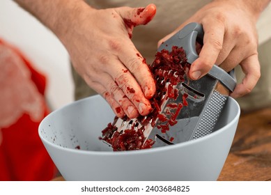 Close-up of hands with red stains using a grater to shred beetroot into a white bowl, healthy cooking concept.