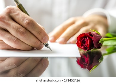 Closeup of the hands of a romantic man writing a love letter to his sweetheart for Valentines Day to be accompanied by a single beautiful fresh red rose