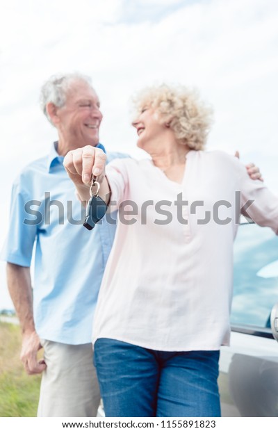 Close-up of the hands of a senior woman showing\
the keys of her car while posing next to her partner as confident\
elderly drivers