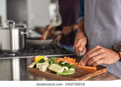 Closeup of hands slicing carrots on chopping board. Closeup of hands cutting vegetables in kitchen near the burners. Detail of man wearing apron chopping vegetables for a recipe at home.
