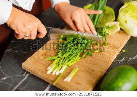 Close-up of hands slicing green onions on a wooden cutting board with a kitchen knife.
Fresh ingredients like lettuce and avocado in the background create a healthy cooking atmosphere.