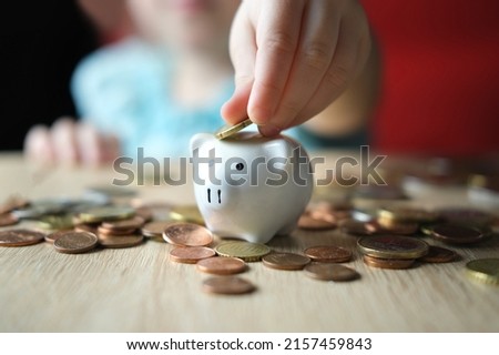 close-up of hands of small child manipulate with metal coins, kid counts, puts euro union money in white piggy bank, concept of pocket money, life insurance, save up for gift, foreground focus
