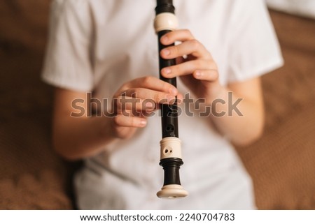 Close-up hands of talented adorable little girl playing flute sitting on bed in bedroom. Child practices playing musical instrument at home. Concept of leisure activity at home.