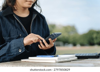 Close-up of hands tapping on back of modern smartphone with triple camera, notebooks and laptop keyboard visible on outdoor stone table in sunlight.
