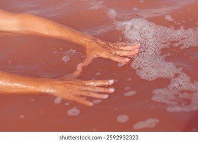 Close-up of hands touching the surface of pink saltwater with bubbles forming around, capturing the serene and tactile moment in a natural salt lake under sunlight.