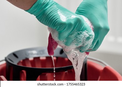 Close-up of hands with turquoise gloves holding and wringing out pink microfiber rag or cleaning cloth. Water and soap sud dripping in red bucket. Hygiene concept.