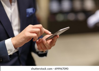 Close-up of hands of unidentified successful man in formal suit typing text in smart phone against background of  blurry interior of fashion boutique - Powered by Shutterstock