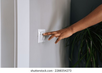Close-up hands of unrecognizable African American woman turning on light in dark room by pushing switch on white wall. Closeup cropped shot of black female turning electricity light switch on wall.