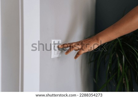 Close-up hands of unrecognizable African American woman turning on light in dark room by pushing switch on white wall. Closeup cropped shot of black female turning electricity light switch on wall.