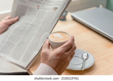 Close-up Of The Hands Of An Unrecognizable Older Man Holding A Newspaper In One Hand And A Cup Of Coffee In The Other, A Closed Laptop On The Table, Concept Of Leisure