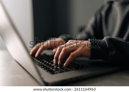 Close-up hands of unrecognizable programmer male typing on laptop keyboard, immersed in software development or coding, sitting at table by window in dark room. Concept of remote job, freelance.