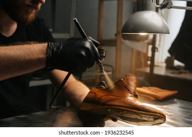 Close-up hands of unrecognizable shoemaker wearing black gloves spraying paint of light brown leather shoes. Concept of cobbler artisan repairing and restoration work in shoe repair shop. - Powered by Shutterstock