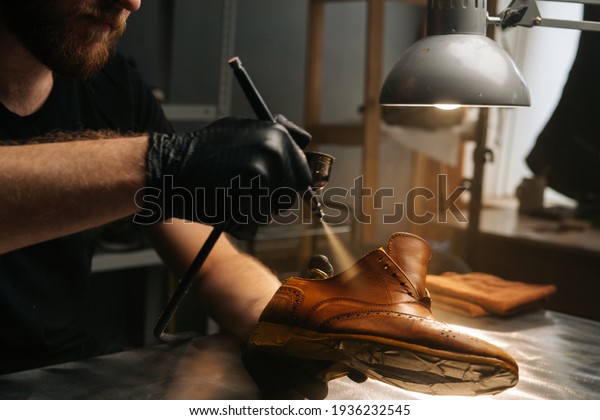 Close-up hands of unrecognizable shoemaker wearing\
black gloves spraying paint of light brown leather shoes. Concept\
of cobbler artisan repairing and restoration work in shoe repair\
shop.