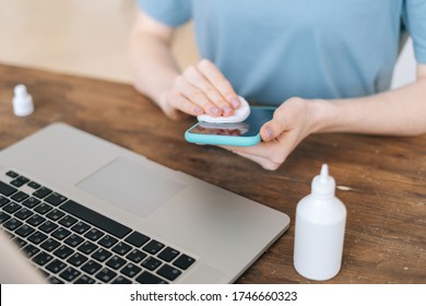 Close-up Of Hands Of Unrecognizable Woman Cleaning Mobile Phone With Antibacterial Sanitizer, Prepares To Starting Work Sitting At The Desk While Sitting At The Table With Laptop At Home Office.