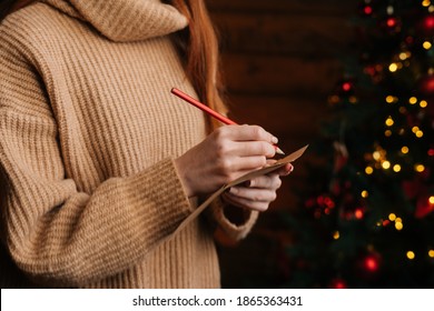 Close-up hands of unrecognizable young woman writing Christmas letter to Santa Claus at cozy dark living room. Xmas tree baubles and tinsel lights blinking at New Year Eve on blur bokeh background.