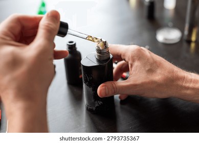 Close-up of hands using a dropper to fill a vape mod with e-liquid on a dark table, surrounded by bottles and vaping accessories.