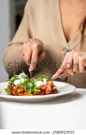 A close-up of a person’s hands using a fork and knife to cut into a beautifully plated dish with fresh vegetables, poached eggs, and herbs, set on a white tablecloth. Elegant dining scene.