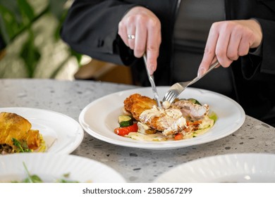 A close-up of hands using a knife and fork to cut into a plated chicken dish with colorful vegetables. The setting is a stylish dining environment, enhancing the culinary experience.