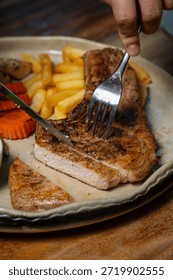 Close-up of hands using knife and fork to slice a grilled pork chop steak served with French fries, corn, carrots, green beans and brown gravy on a ceramic plate in a restaurant.