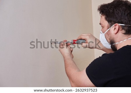 Close-up of hands using a manual screwdriver to install a screw into a wall anchor. Person wearing safety glasses and mask during home repair or DIY project.