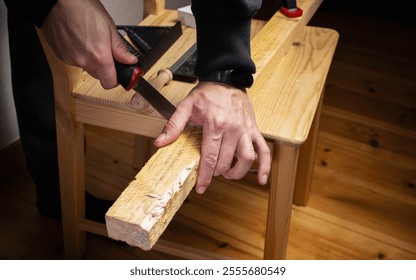 Close-up of hands using a red-handled handsaw to cut a wooden plank on a chair, surrounded by carpentry tools. Perfect for woodworking, DIY, or crafting themes. Horizontal composition - Powered by Shutterstock