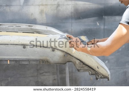 Close-up of hands using sanding tools to smooth primer and body filler on a car bumper before painting.