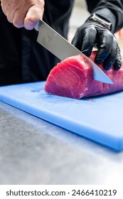 Close-up of hands wearing black gloves slicing red tuna fish on a blue cutting board with a metal knife, depicting food preparation