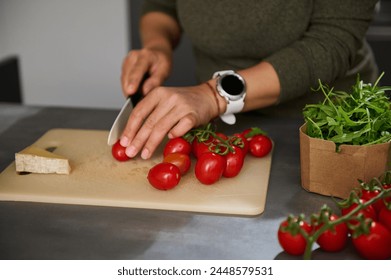 Close-up hands of a woman chopping cherry tomato on a cutting board, preparing healthy salad with fresh arugula leaves, standing at kitchen counter. Culinary, diet, healthy eating and food concept - Powered by Shutterstock