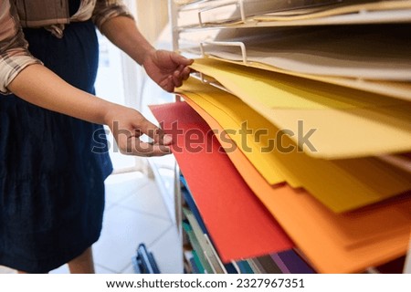 Close-up hands of a woman, customer in creative art store, choosing design paper on shelves with colorful papers for watercolor painting, pastel drawing, fine art. Creativity. Hobby. Occupation