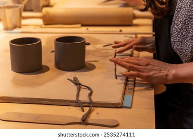 Close-up of hands working with clay, shaping cylindrical pots in a ceramic workshop. The warm tones of the setting emphasize creativity and craftsmanship in pottery making. - Powered by Shutterstock