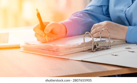 Close-up of hands writing in a notebook with a ring binder. Person wears a blue shirt and holds a pen. Warm sunlight illuminates the wooden table. Focused action suggests concentration