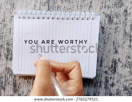 Close-up of hands writing “You Are Worthy” in a notebook, symbolizing self worth, motivation, encouragement, mental wellness, and positive affirmation.