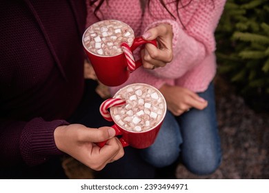 Close-up of hands of young couple holding red cups with hot cocoa, marshmallows, striped red, white candy cane. Man and woman are having fun at Christmas tree market. Faceless view. Copy space - Powered by Shutterstock