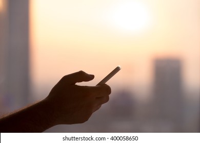 Close-up Of Hands Of Young Man Holding Mobile Phone, Using Smartphone App, Scrolling. Silhouette Against Sunny Street View