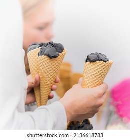 Close-up of Hands of young man with three trendy Delicious fresh charcoal ice cream in waffle cone for himself and for friends, blurred unrecognizable silhouette of girl