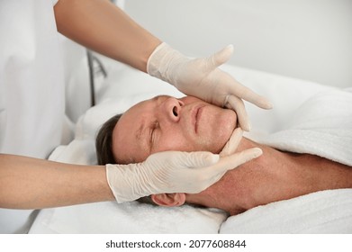 Close-up Of Handsome Mature 50 Years Old Man Relaxing On Massage Table While Getting Professional Facial Lifting Massage At Spa. Anti-aging, Rejuvenation, Face Lifting And Skin Care Procedures Concept