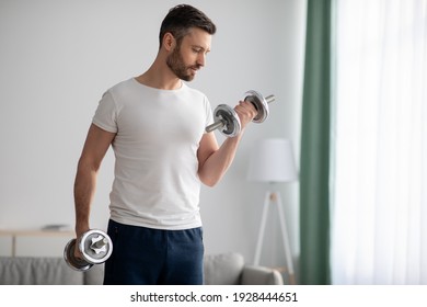 Closeup of handsome middle-aged man doing dumbbell workout at home, working on arms strength, looking at his biceps, copy space. Athletic man lifting dumbells up over living room interior - Shutterstock ID 1928444651