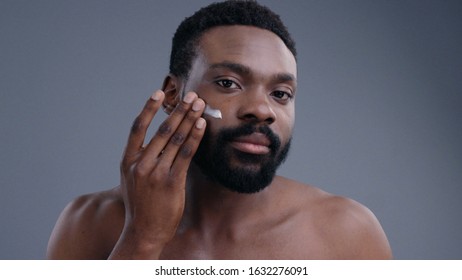 Close-up Of Handsome Sensitive Young Black Man Applying Facial Cream Looking At Camera On Grey Background. Male Beauty And Care Concept.