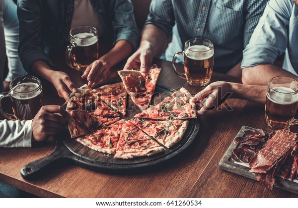 Close-up of handsome young friends drinks beer and\
eating pizza at pub.