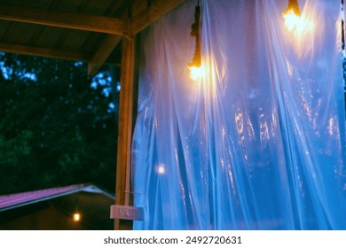 Close-up of hanging light bulbs inside a wooden structure with transparent plastic sheets, set against a backdrop of a lush, dark forest. - Powered by Shutterstock