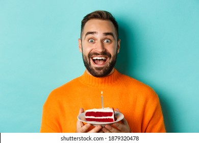 Close-up of happy adult man celebrating birthday, holding bday cake with candle and making wish, standing against turquoise background