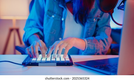 Close-up happy asia girl dj use launchpad synthesizer keyboard sound mixer wear headphone made music in laptop computer online live in living room home studio at night. Content creator concept.