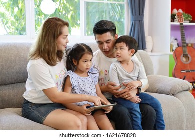 closeup happy Asian family teaching their children son and daughter how to use tablet while sitting on grey sofa in living room with smiling faces (family and technology concept)