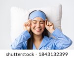 Close-up of happy beautiful asian girl in sleeping mask and blue pajamas lying in bed, smiling upbeat, having perfect mood as going bed and getting ready sleep, standing white background