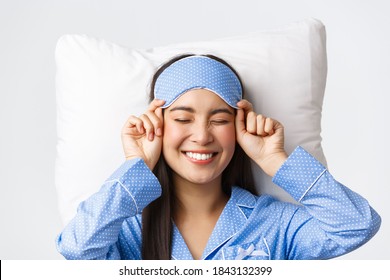 Close-up of happy beautiful asian girl in sleeping mask and blue pajamas lying in bed, smiling upbeat, having perfect mood as going bed and getting ready sleep, standing white background